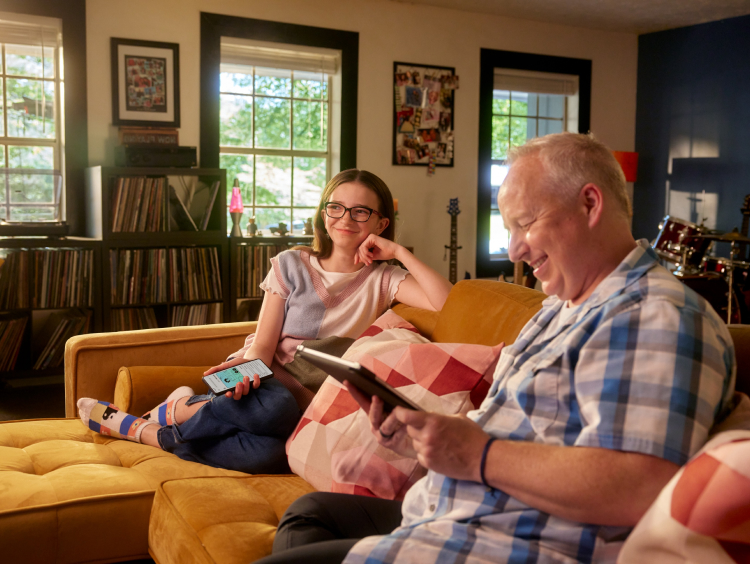 A girl and her dad sit on the couch while her dad smiles at a tablet powered by Brightspeed DSL internet.