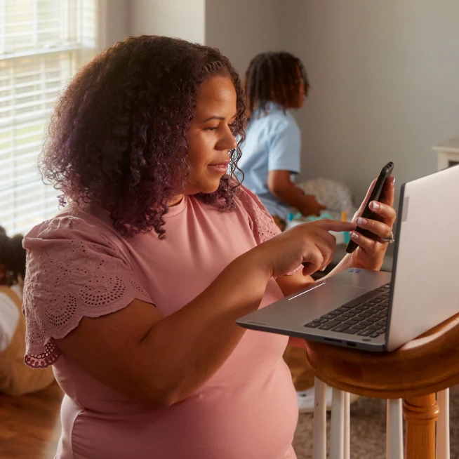 A lady is doing WFH, standing in front of her laptop and browsing Moblie powered by Brightspeed Internet.