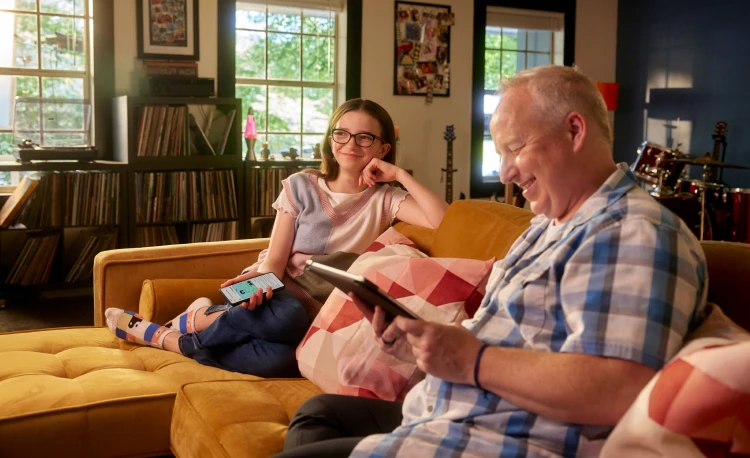 A girl and her dad sit on the couch while her dad smiles at a tablet.