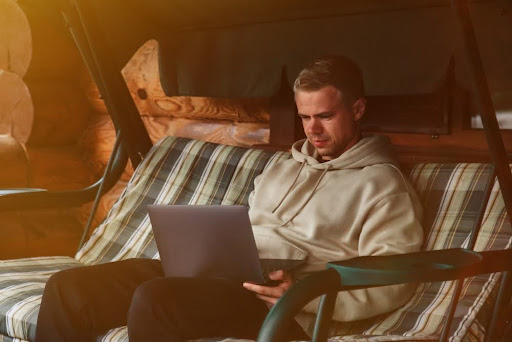 A man in a beige hoodie sits on a striped outdoor swing, focused on his laptop. 