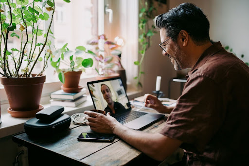 Man in brown shirt having a video call on a laptop at a wooden desk, surrounded by potted plants and closed windows.