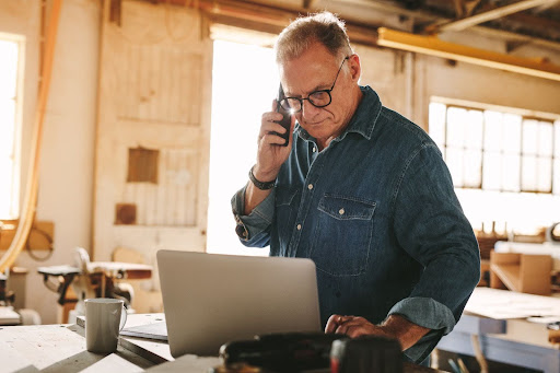 Middle-aged man in denim shirt talks on phone, works on laptop in a woodworking shop.