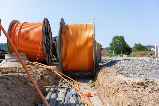 Large spools of orange cables rest beside a trench at a construction site under a clear sky, with distant trees and houses in the background.