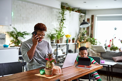 A woman works on a laptop and talks on the phone at a table, while a child in pajamas uses a tablet.