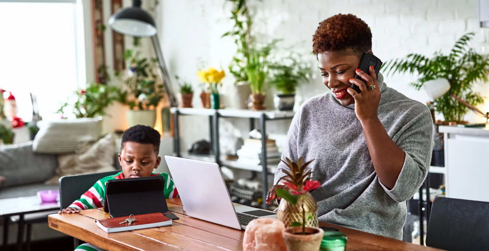 A mother sitting at a the kitchen table looking talking on the phone and browsing on her laptop, while her child is sitting next to her looking at a tablet.
