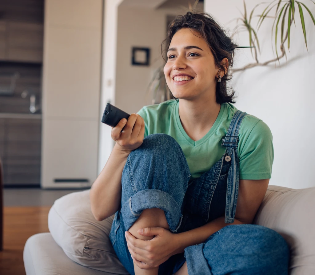 A young woman sitting on the couch and smiling while pointing a remote control to the tv.