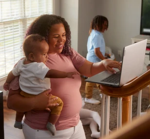 A woman holding a baby in one arm while using a laptop connected to Brightspeed Fiber Internet with the other hand.