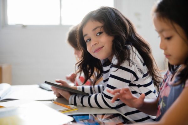 Several children sitting at a table while using tablet devices in a brightly lit room.