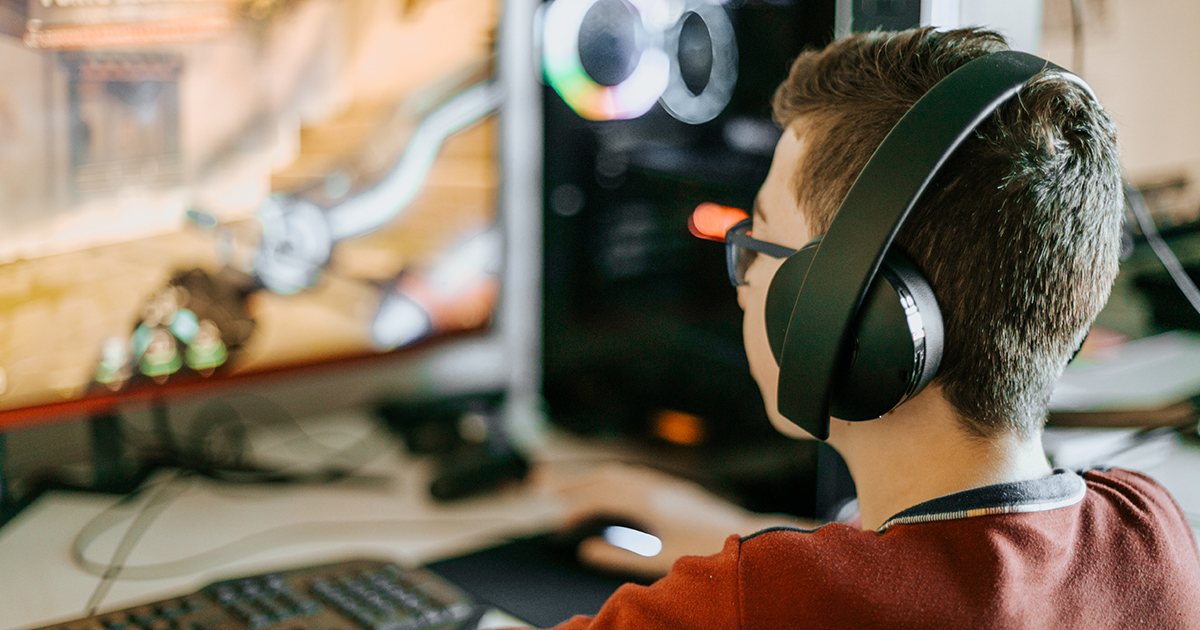 A teenager playing a game on his desktop computer while wearing headphones