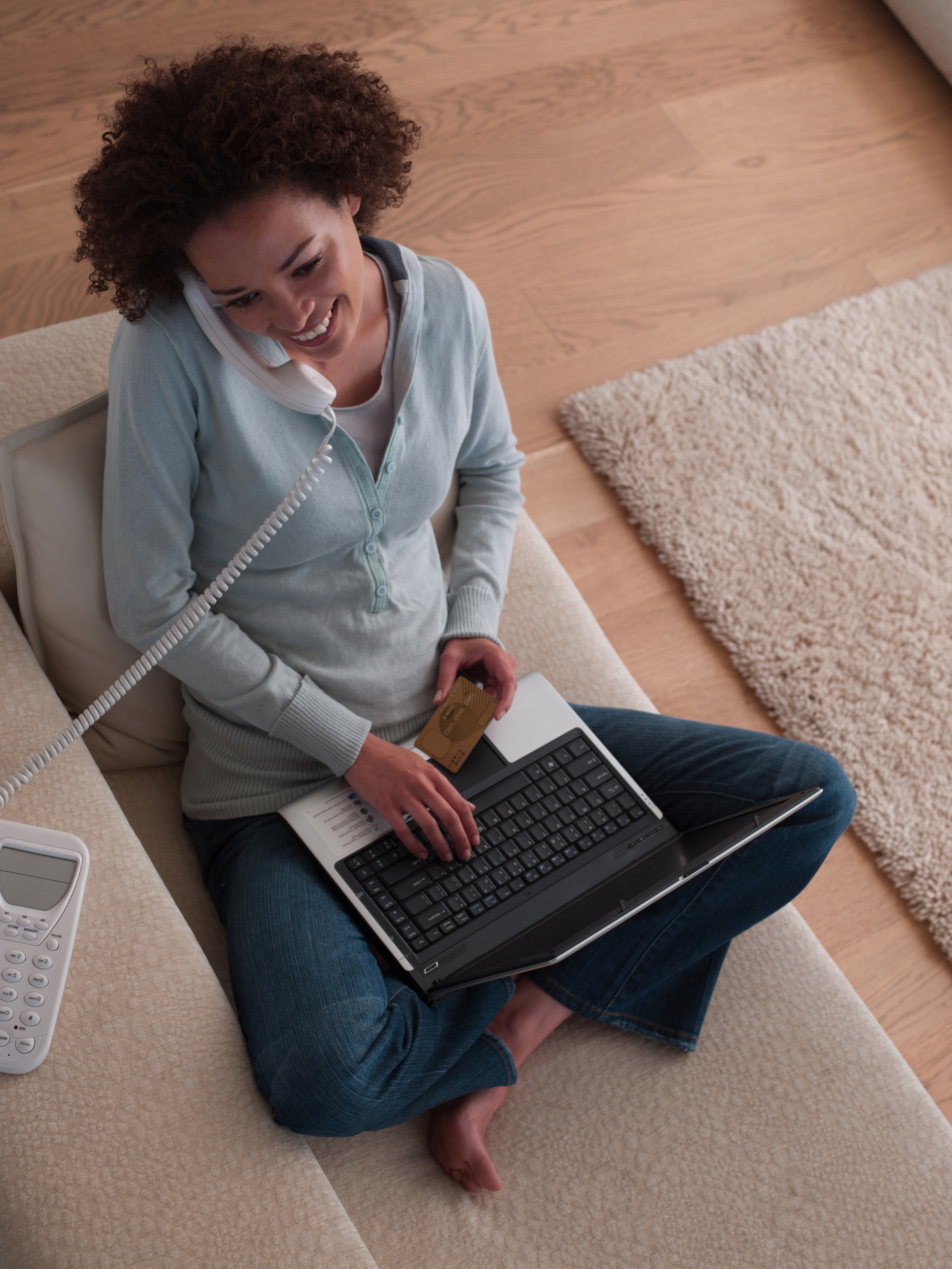 A woman sitting on her couch while on her phone, holding a credit card and using her laptop.